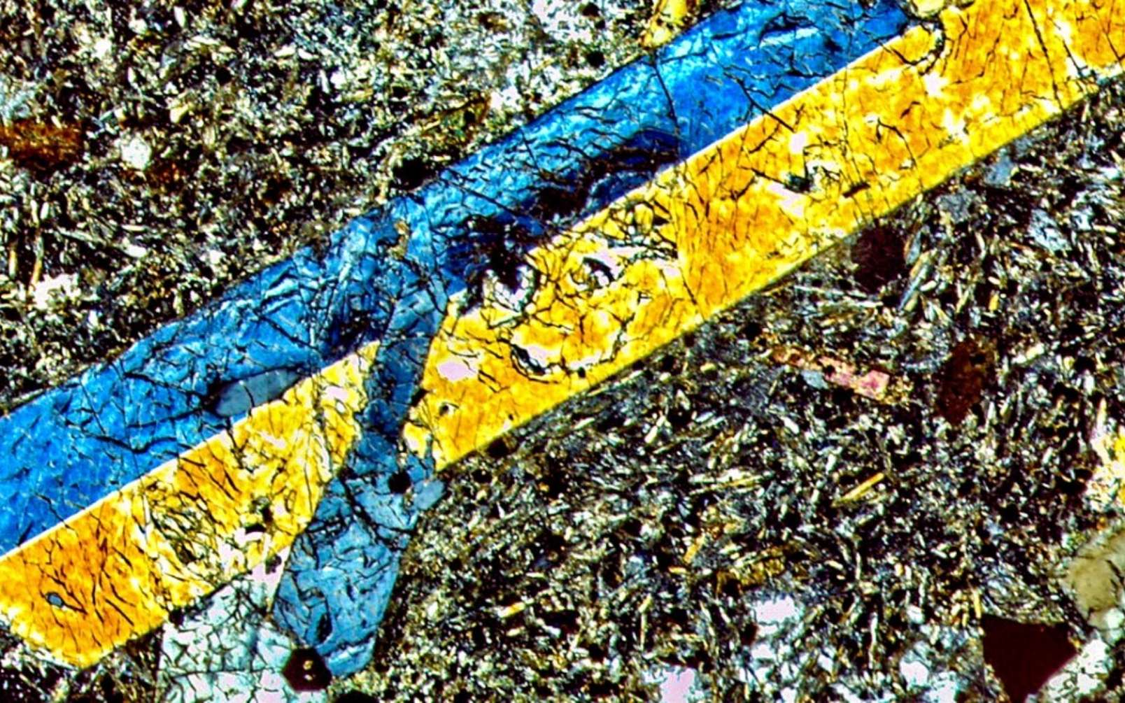 Dans cette image en lumière polarisée, on observe au microscope un échantillon de lave provenant des Bermudes. Les cristaux bleus et jaunes sont constitués d'augite de titane, entourés d'une masse de minéraux, qui comprend feldspaths, phlogopite, spinelle, pérovskite et de l'apatite. Cet assemblage suggère que la source du manteau qui a produit cette lave était riche en eau. © Gazel Lab