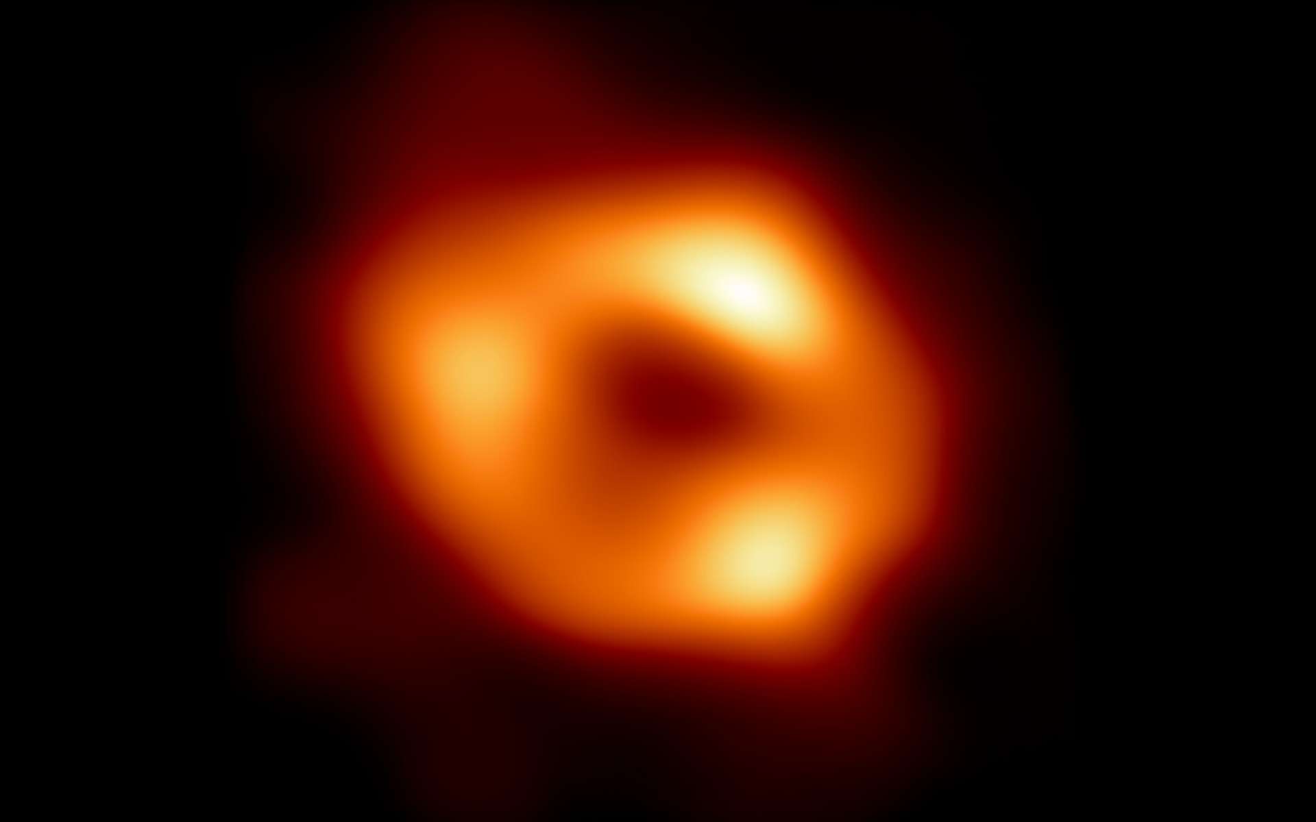 Voici la première image de Sgr A*, le trou noir supermassif au centre de notre galaxie, avec un fond noir ajouté pour s'adapter à des écrans plus larges. Il s'agit de la première preuve visuelle directe de la présence de ce trou noir. Elle a été obtenue par l'Event Horizon Telescope (EHT), un réseau qui relie huit observatoires radio existants sur la planète pour former un seul télescope virtuel de la taille de la Terre. Le télescope porte le nom de l'horizon des événements, la limite du trou noir au-delà de laquelle aucune lumière ne peut s'échapper. © EHT Collaboration