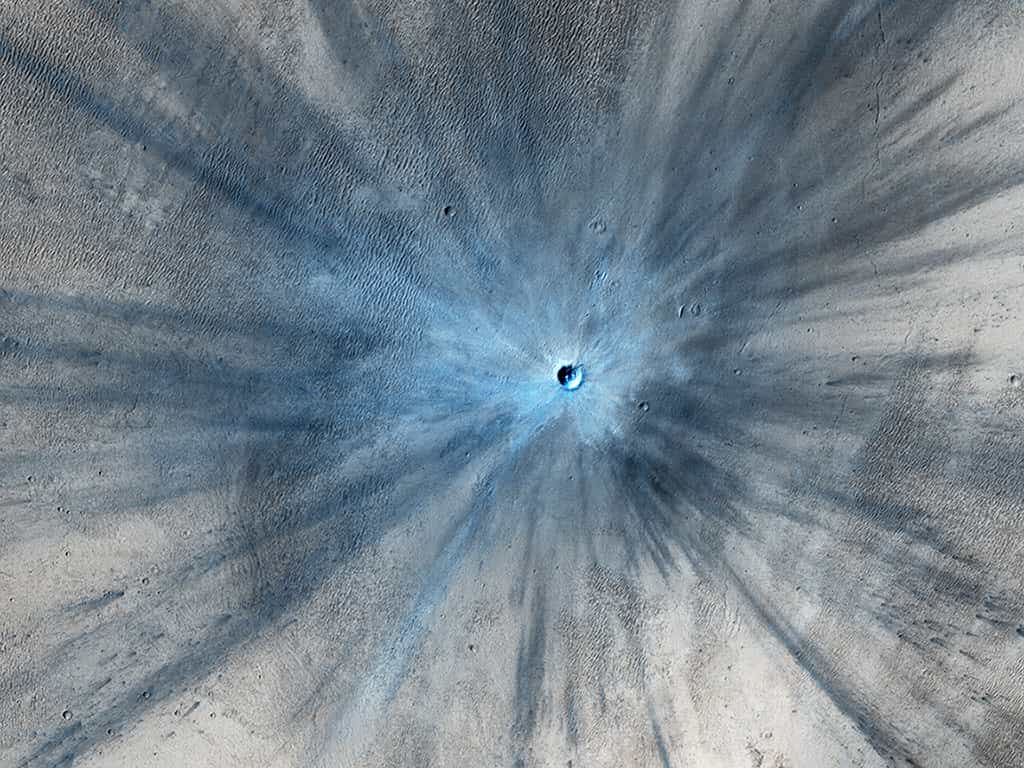 Jeune cratère rayonnant de 30 mètres de diamètre photographié le 19 novembre 2013 par la sonde spatiale Mars Reconnaissance Orbiter (MRO). La caméra HiRise révèle en fausses couleurs les éjectas de poussières rouges (ici en bleu) qui s'étalent dans un rayon de 15 km autour de l'impact. © Nasa, JPL, University of Arizona