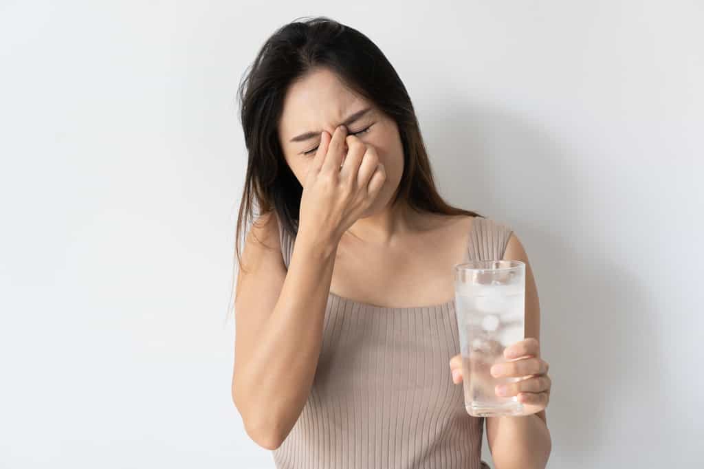 Jeune femme ressentant un terrible mal de tête ou « brain freeze » après avoir bu un verre d'eau glacé. © Pattarisara, Adobe Stock