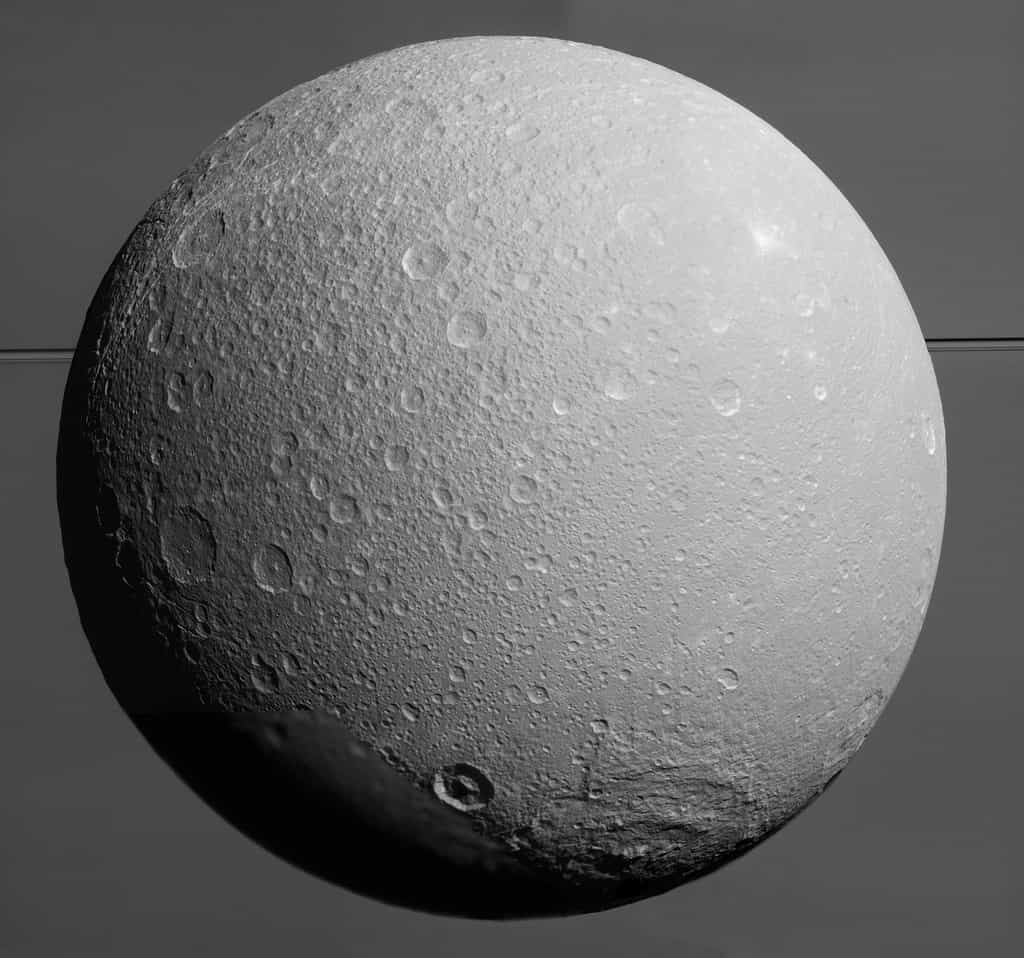 Au premier-plan, Dioné et sa surface grêlée de cratères d’impact et à l’arrière-plan, on distingue l’atmosphère de Saturne et ses anneaux. L’image a été prise par Cassini le 17 août 2015, à 170.000&nbsp;km de sa surface lorsqu’elle entreprenait le dernier survol de sa mission de cette lune glacée. © Nasa, JPL-Caltech, Space Science Institute