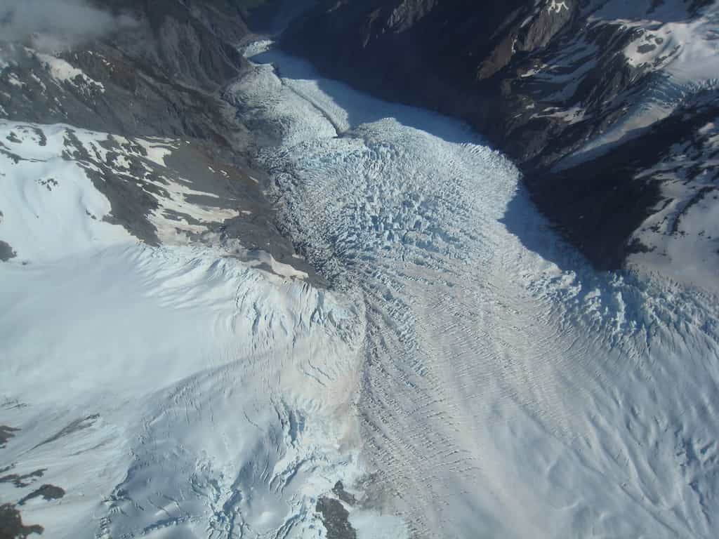 Vue aérienne du glacier Franz Josef qui s’étend sur environ 10 km et glisse à une vitesse pouvant atteindre 2 m/j sur un substrat rocheux composé de schistes métamorphiques. Ces schistes contiennent les matériaux graphitiques utilisés pour tracer la provenance des particules sédimentaires dans le ruisseau sous-glaciaire. © Avenue, Wikimedia Commons, CC BY-SA 3.0