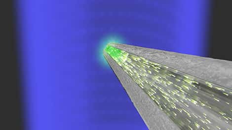 Schéma du dispositif. Le rayonnement bleu représente les rayons X ; la sphère verte intense et brillante correspond au fragment de scintillateur ; les étincelles à l'intérieur du corps de l'antenne optique symbolisent l'émission de photons par l'agrégat luminescent sous l'effet des rayons X. Grâce à l'antenne optique, cette émission de lumière est fortement dirigée vers une fibre optique monomode très étroite. © Miguel Angel Suarez, FEMTO-ST (CNRS, université de Franche-Comté, UTMB, ENSMM)