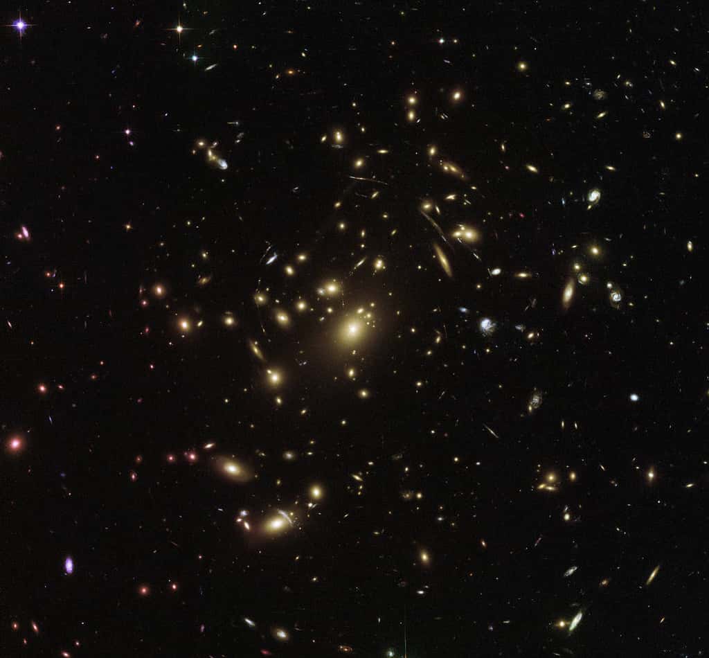 Les amas, comme ici Abell 2537 observé par Hubble, produisent un effet de lentille gravitationnel, c’est-à-dire qu’ils courbent l’espace-temps et nous permettent de voir la lumière de galaxies très lointaines, cachées derrière eux, visibles sur cette image sous forme de traits lumineux. © ESA, Hubble, Nasa