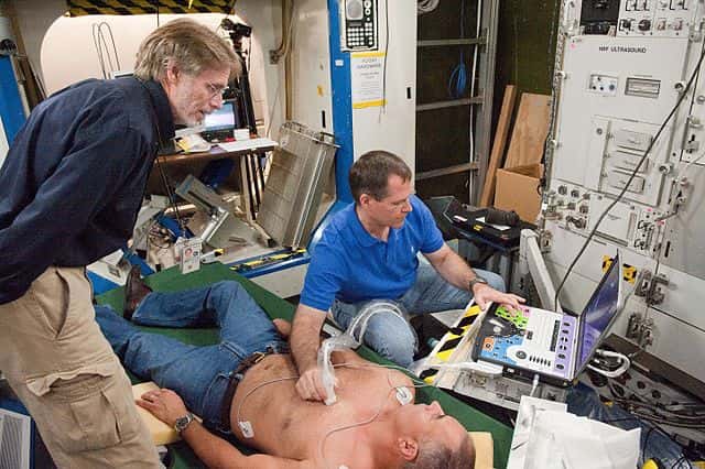 Dans le cadre du programme de suivi par ultrasons, les astronautes sont entraînés pour manipuler correctement l’appareil. Ils se scannent mutuellement le dos, mais pas seulement : les ultrasons permettent aussi d’analyser les muscles, la circulation sanguine et beaucoup d’autres fonctions physiologiques. © Nasa, <em>Wikimedia Commons</em>