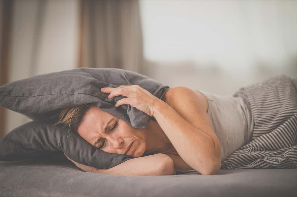 (Une femme couchée dans un lit se bouche les oreilles avec son oreiller.) Le Hum est souvent une source de fatigue et de stress pour ses entendeurs. © lightpoet, Adobe Stock