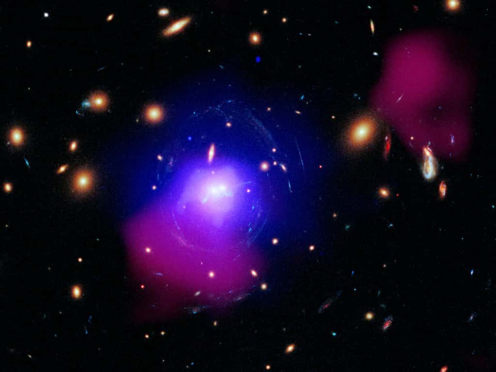 Il s'agit d'une image d'un amas de galaxies appelé SDSS J1531+3414 en rayons X, dans le visible et en radio. La scène globale ressemble à un affichage coloré de lumières comme si elle était vue à travers une fenêtre en verre mouillée. Des points orange flous de différentes tailles sont dispersés sur un fond noir. Ces points orange représentent des galaxies entières. Près du centre de l’image, deux galaxies centrales apparaissent sous forme de points blancs brillants. Des amas d'étoiles, ressemblant à des perles sur un fil dans des tons de bleu électrique s'étendent à travers l'espace à partir des deux galaxies, puis s'enroulent légèrement. Des nuages ​​de lumière bleue, des rayons X, et de lumière radio rose foncé, entourent les deux galaxies. © Rayons X : Nasa/CXC/SAO/O. Omoruyi et coll.; Visible : Nasa/ESA/STScI/G. Tremblay et coll.; Radio : Astron/Lofar ; Traitement d'images : Nasa/CXC/SAO/N. Wolk
