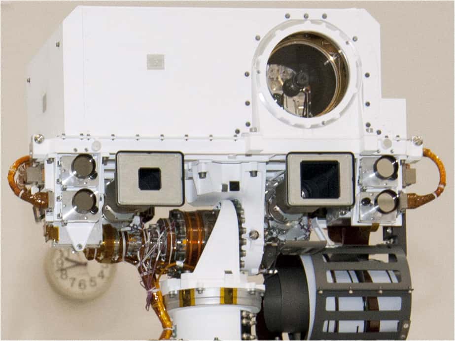 En haut du mât de Curiosity, l'instrument ChemCam (la boîte blanche avec la grosse lentille) envoie un laser pulsé sur une cible tandis que le capteur, derrière l'objectif, filme le nuage de plasma lumineux produit par cet échauffement. Un spectromètre analyse ensuite l'émission de cette lueur pour déterminer les composants présents dans la roche. On remarque les objectifs à ouverture carrée des deux caméras de mât, avec une focale longue à gauche et courte à droite. © Nasa, JPL-CalTech