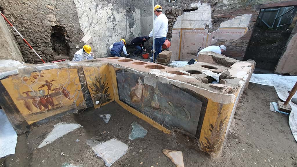 Vue d'ensemble du thermopolium complètement excavé à Pompéi. On voit la fresque représentant la néréide et celle de l'homme portant des charges. © Site historique de Pompéi