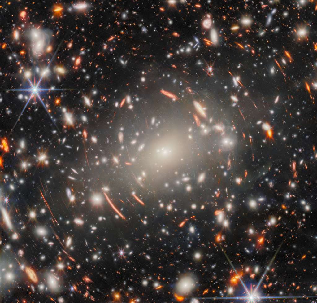 Un champ de galaxies dans l'espace, dominé par une énorme galaxie elliptique d'un blanc éclatant, cœur d'un amas de galaxies massif. De nombreuses autres galaxies elliptiques sont visibles autour. Autour d'elle se dessinent également de courtes lignes rouges incurvées et brillantes, images de galaxies lointaines en arrière-plan, agrandies et déformées par l'effet de lentille gravitationnelle. Deux étoiles au premier plan apparaissent grandes et brillantes, entourées de longues pointes de diffraction. © ESA/Webb, Nasa et ASC, H. Atek, M. Zamani Un champ de galaxies dans l'espace, dominé par une énorme galaxie elliptique d'un blanc éclatant, cœur d'un amas de galaxies massif. De nombreuses autres galaxies elliptiques sont visibles autour. Autour d'elle se dessinent également de courtes lignes rouges incurvées et brillantes, images de galaxies lointaines en arrière-plan, agrandies et déformées par l'effet de lentille gravitationnelle. Deux étoiles au premier plan apparaissent grandes et brillantes, entourées de longues pointes de diffraction. © ESA/Webb, Nasa et ASC, H. Atek, M. Zamani
