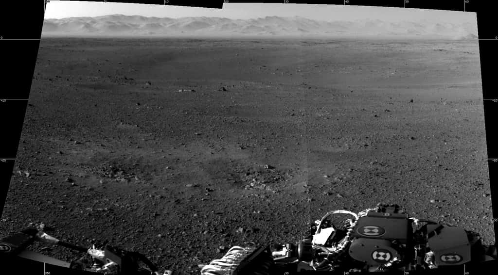 La première image en haute résolution du site d'atterrissage de Curiosity, juxtaposition de deux photographies prises avec les « navcams », caméras de navigation, installées en haut du mât, lequel a été correctement redressé. À l'horizon se dresse le rebord du cratère Gale, dont l'aspect irrégulier est dû à la longue érosion qu'il a subie. Les deux dépressions visibles devant le robot ont été creusées par le souffle des rétrofusées de la grue aéroportée. © Nasa/JPL-Caltech
