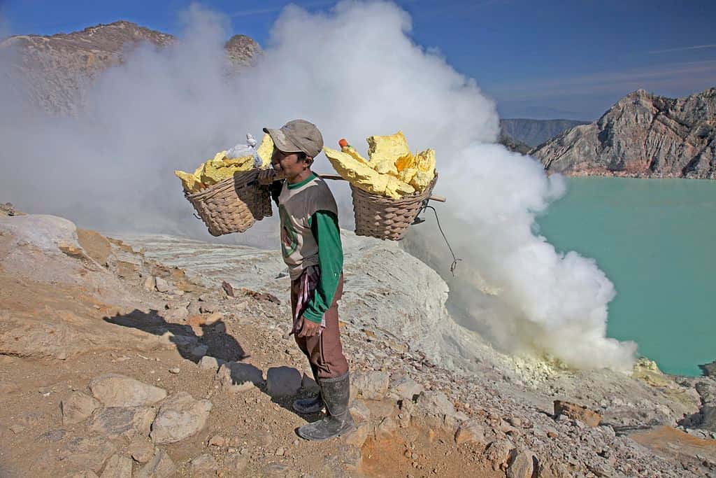 Cet homme porte de grandes quantités de minerais de soufre extraites du solfatare. Derrière, on peut observer le lac du cratère, considéré comme le plus acide au monde. © Jean-Marie Hullot, Wikipédia, cc by sa 2.0