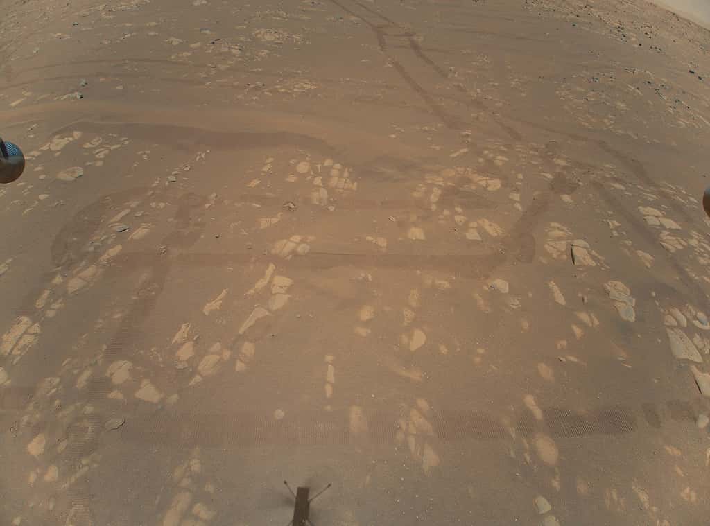 Sur cette première image en couleur renvoyée par Ingenuity, l’ombre de l’hélicoptère à proximité du site de décollage et les traces laissées au sol par le rover Perseverance. © Nasa, JPL-Caltech
