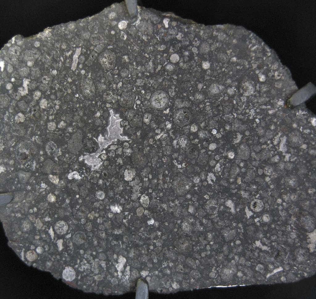 Une coupe de la météorite d'Allende. Chaque cercle blanc, gris ou beige représente la section d'un chondre. Ces chondres sont inclus dans une matrice (mélange de silicates et de fer), ici de couleur gris foncé. Cette chondrite carbonée contient également des structures irrégulières blanchâtres, des CAI (pour calcium-aluminum inclusions, en anglais). Leur formation date de 4,568 milliards d'années et elles contiennent des traces de radioactivités éteintes. © <em>Wikipedia-Shiny Things</em>, Pierre Thomas 