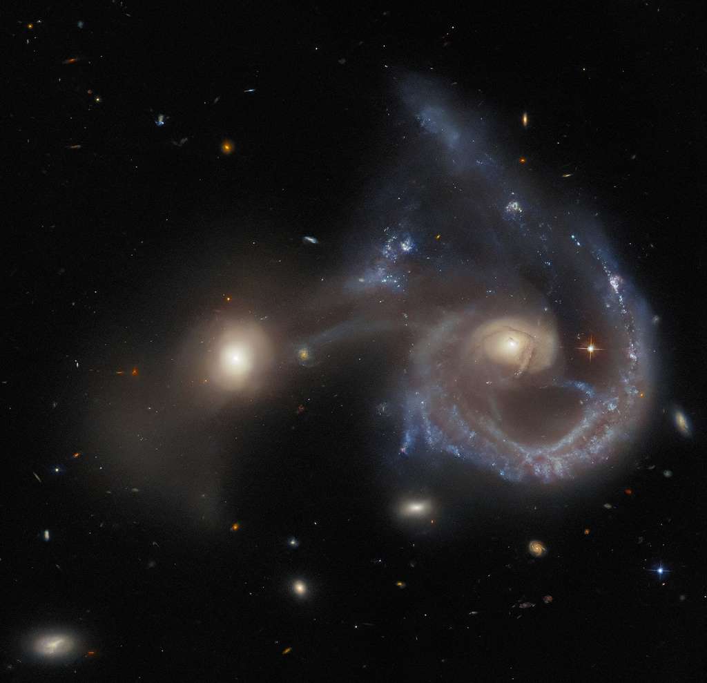 Cette image prise par Hubble avec la caméra avancée ACS (<em>Advanced Camera for Surveys</em>) du télescope spatial Hubble de la Nasa/ESA montre Arp 107, un objet céleste composé d’une paire de galaxies en pleine collision. La plus grande galaxie (à gauche de l'image) est une galaxie extrêmement énergétique d’un type connu sous le nom de galaxie de Seyfert, qui abrite des noyaux galactiques actifs dans leur cœur. Les galaxies de Seyfert sont remarquables car, malgré l’immense luminosité de leur noyau actif, le rayonnement de toute la galaxie peut être observé. Cela est évident sur cette image, où les spirales de toute la galaxie sont facilement visibles. La plus petite est reliée à la plus grande par un « pont » apparemment ténu, composé de poussière et de gaz. Arp 107 fait partie d’un catalogue de 338 galaxies connu sous le nom d’Atlas des galaxies particulières, compilé en 1966 par Halton Arp. Elle a été observée par Hubble dans le cadre d’un programme d’observation qui cherchait spécifiquement à combler une « lacune » en matière d’observation, en effectuant des observations limitées des membres du catalogue Arp. L’objectif était en partie de fournir au public des images de ces galaxies spectaculaires et difficiles à définir. © ESA-Hubble & Nasa, J. Dalcanton, Dark Energy Survey/DOE/FNAL/NOIRLab/NSF/AURA, SDSS 