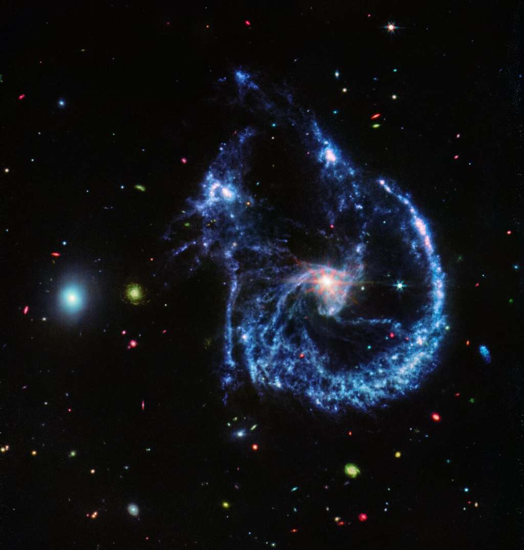 Cette image d’Arp 107 en fausses couleurs, obtenue par l’instrument Miri (<em>Mid-InfraRed Instrument</em>) de Webb, révèle le trou noir supermassif qui se trouve au centre de la grande galaxie spirale à droite, comme en témoigne le petit « noyau » central brillant. Ce noyau brillant, où le trou noir attire une grande partie de la poussière dans des couloirs, présente également les pics de diffraction caractéristiques de Webb, causés par la lumière qu’il émet en interaction avec la structure du télescope lui-même. La caractéristique déterminante de la région, révélée par Miri, est peut-être les millions de jeunes étoiles qui se forment, surlignées en bleu. Ces étoiles sont entourées de silicates poussiéreux et de molécules semblables à de la suie, connues sous le nom d’hydrocarbures aromatiques polycycliques. La petite galaxie elliptique à gauche, qui a déjà achevé une grande partie de sa formation d’étoiles, est composée de nombreuses de ces molécules organiques. © Nasa, ESA, CSA, STScI