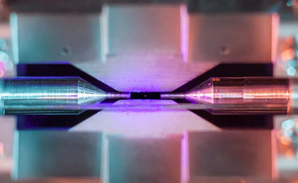 La distance entre les pointes d'aiguille est d'environ deux millimètres. Lorsqu'il est éclairé par un laser de couleur bleue-violette, l'ion de strontium piégé dans l'expérience absorbe et réémet des photons rapidement. Avec un appareil photo ordinaire, une pause longue accumule ces grains de lumière en observant à travers une fenêtre de la chambre à ultra-vide qui abrite le piège à ions. La petite tache colorée qui apparaît alors ne montre bien sûr pas la taille de l'atome mais elle signale spectaculairement sa présence. © David Nadlinger, <em>University of Oxford</em>