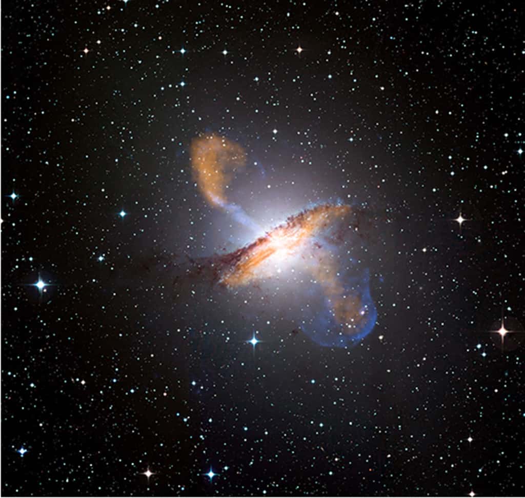 La galaxie elliptique NGC 5128, qui est aussi une puissante radiosource : Centaurus A. En blanc se trouve la galaxie optique (les étoiles, vues avec un télescope de l’ESO), et l’obscuration due à la poussière en noir ; en orange, l’émission de la poussière en ondes submillimétriques (télescope Apex, <em>Atacama Pathfinder Experiment</em>), en bleu l’émission des rayons X (satellite Chandra). © Chandra, Nasa