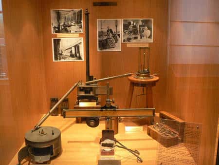 Le « montage Curie » de mesure de la radioactivité (exposé au musée Curie à Paris) : à gauche la « boîte » est la chambre d’ionisation où est placé l’échantillon dont on mesure l’activité, au centre (au fond) l’électromètre, au premier plan, la réglette graduée et, accrochée dessous la lampe dont la lumière est réfléchie par le miroir suspendu au fil de l’électromètre. À droite, au fond, la balance à quartz ajoutée ensuite, et sa boîte de poids.