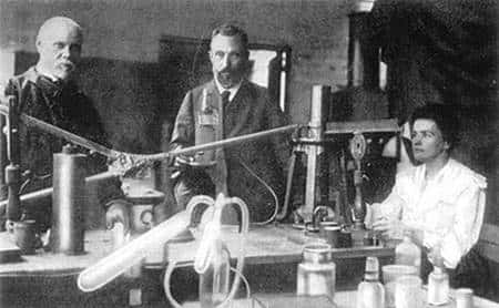 Marie Curie aux commandes, la main droite sur le poids de la balance à quartz, la main gauche sur le chronomètre et l’œil fixé sur la tache de lumière sur la règle. À ses côtés, Pierre Curie et son assistant, Petit. L’électromètre est ici en dehors de l’image, sur la gauche. ©ACJC