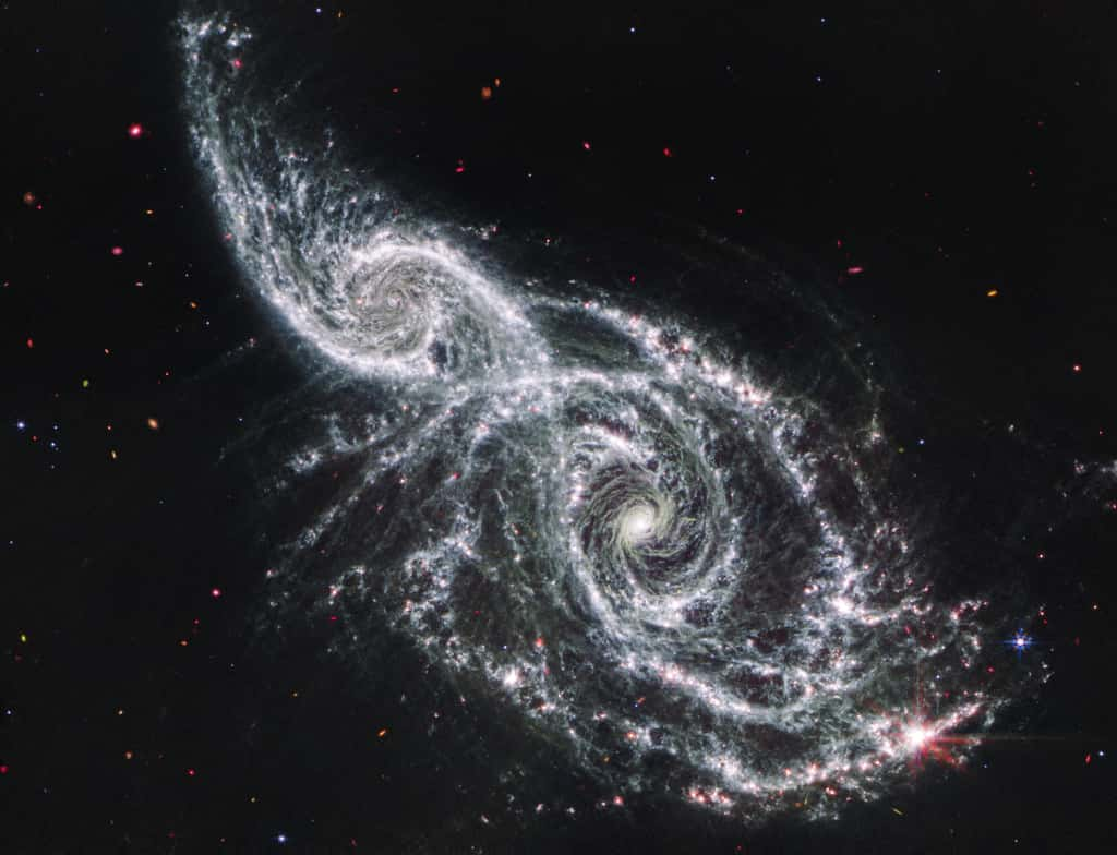 L’image infrarouge moyenne des galaxies IC 2163 et NGC 2207 prise par le télescope spatial James-Webb rappelle la froideur des ossements morts depuis longtemps mêlés à des vapeurs étranges. Deux grands « yeux » lumineux se trouvent au cœur des galaxies, et des bras spiraux vaporeux s’étendent dans les vastes distances de l’espace. L’image infrarouge moyenne de Webb excelle à montrer où la poussière froide brille dans ces galaxies – et permet de localiser les étoiles et les amas d’étoiles enfouis dans la poussière. Trouvez ces régions en recherchant les points roses le long des bras spiraux. Beaucoup de ces zones abritent des étoiles en formation active qui sont encore enfermées dans le gaz et la poussière qui alimentent leur croissance. D’autres points roses peuvent être des objets qui se trouvent bien derrière ces galaxies, notamment des trous noirs supermassifs actifs extrêmement éloignés connus sous le nom de quasars. La région rose la plus grande et la plus brillante, qui scintille avec huit pics de diffraction proéminents en bas à droite, est une mini-explosion d'étoiles, un endroit où de nombreuses étoiles se forment en succession rapide. Trouvez les trous en forme de dentelle dans les bras spiraux. Ces zones regorgent de formations d'étoiles. Enfin, scannez le fond noir de l'espace, où les objets brillent de mille feux dans un arc-en-ciel de couleurs. Les cercles bleus avec de minuscules pics de diffraction sont des étoiles au premier plan. Les objets sans pics sont des galaxies très lointaines. © Nasa, ESA, CSA, STScI