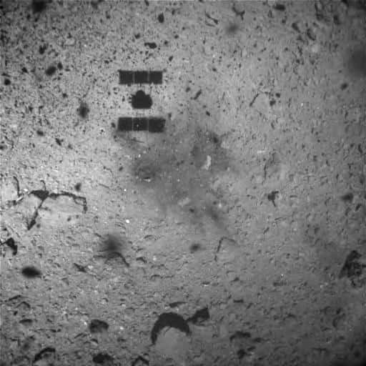 Image de la surface de Ryugu capturée une minute après le <em>touchdown</em> du 22 février 2019 (heure du Japon) par l'Optical Navigation Camera - Wide Angle (ONC-W1) de Hayabusa-2. La sonde était alors à environ 25 m d'altitude. Une tache est visible sur le sol à côté de son ombre. Cette décoloration de la surface provient peut-être de la matière soufflée par les propulseurs de Hayabusa-2 ou par l'impact de son projectile. © Jaxa, <em>University of Tokyo, Kochi University, Rikkyo University, Nagoya University, Chiba Institute of Technology, Meiji University, University of Aizu</em>, AIST