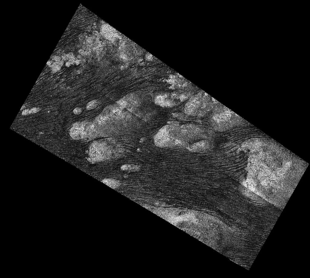 Cette image provient du radar à synthèse d'ouverture (SAR) du vaisseau spatial Cassini de la Nasa et montre la mer de sable de Shangri-La sur Titan. Des centaines de dunes de sable sont visibles sous forme de lignes sombres serpentant à la surface. Ces dunes présentent des motifs d'ondulation et de divergence autour des montagnes élevées (qui apparaissent brillantes au radar), montrant ainsi la direction du vent et du transport du sable à la surface de Titan. © Nasa, JPL-Caltech, ASI
