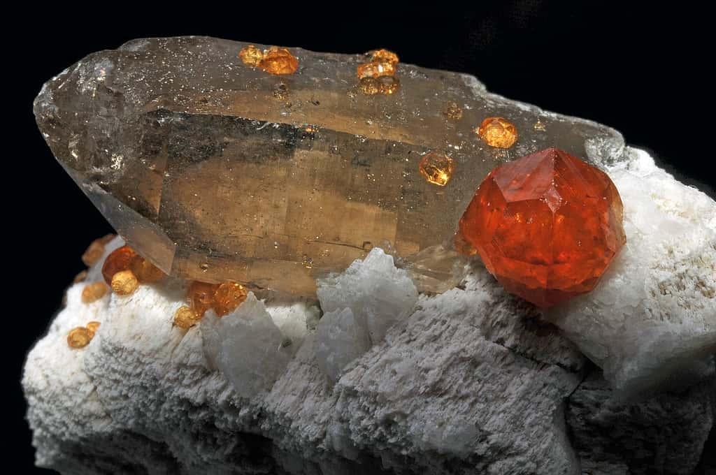 Différents habitus pour différents minéraux ici associés : quartz fumé, grenats (minéraux rouges globuleux) et feldspaths (minéraux blancs). © Géry Parent, <em>Wikimedia Commons</em>, CC by-sa 3.0 