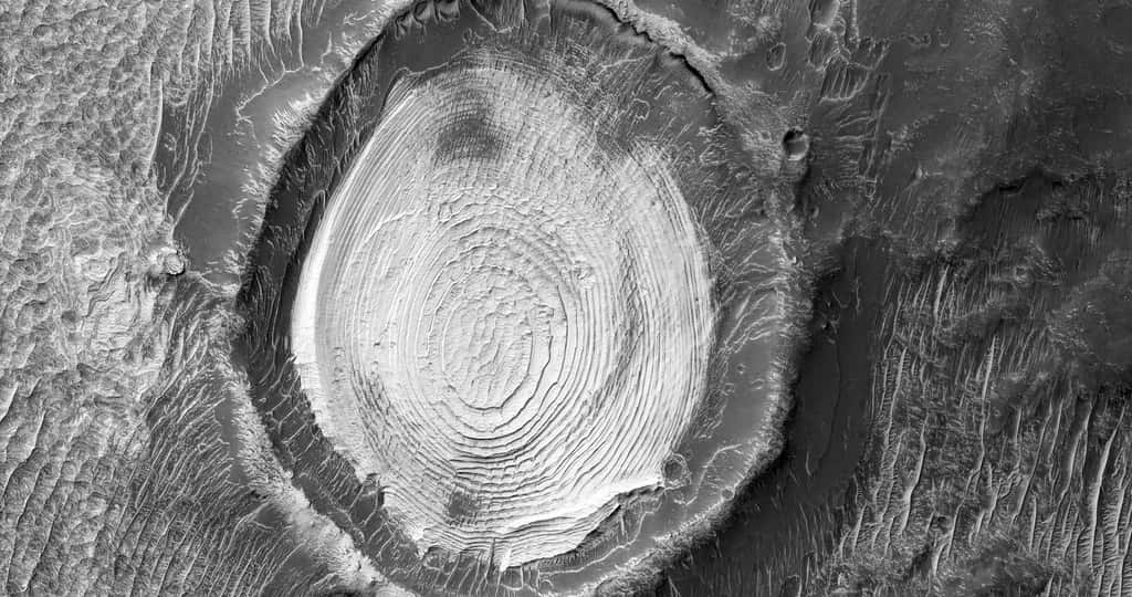 Cette image en noir et blanc montre un cratère dont l’intérieur présente une stratification dont l’origine et la nature ne sont pas déterminées. © Nasa/JPL/UArizona