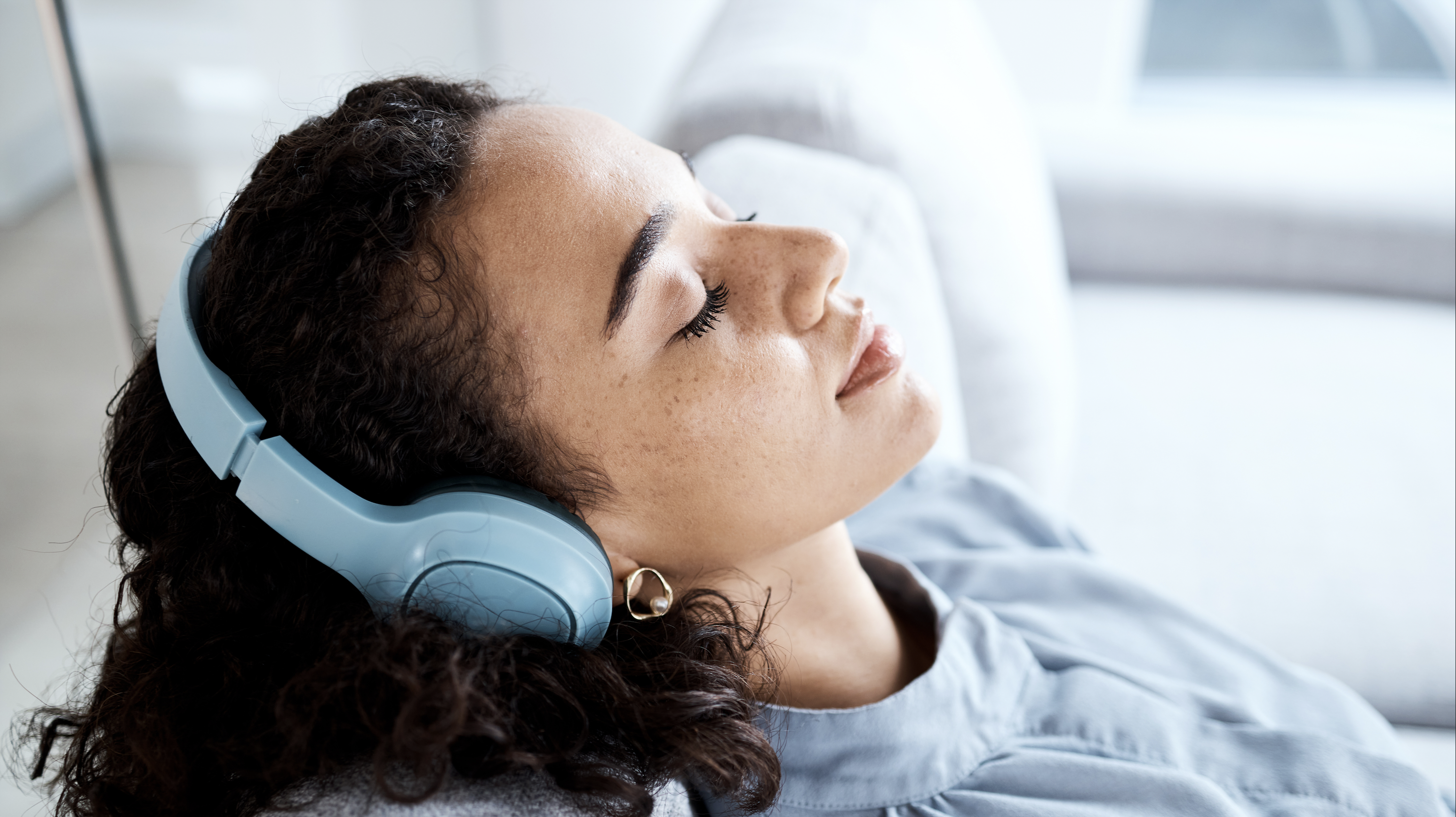 Une femme assise de manière détendue, les yeux fermés et la tête appuyée sur le dossier d'un canapé. Elle porte des écouteurs. © Adobe Stock, Rene La/peopleimages.com