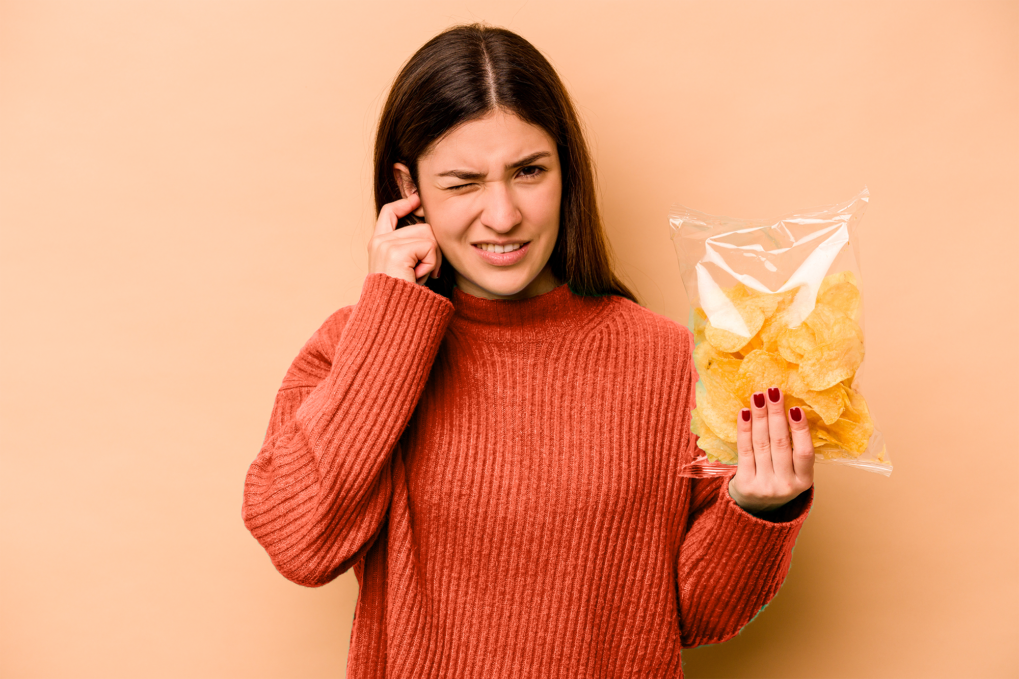 Une jeune femme se bouche une oreille d'une main et tient paquet de chips de l'autre. Son visage indique qu'elle est gênée par le bruit du sachet. © Asier, Adobe Stock   