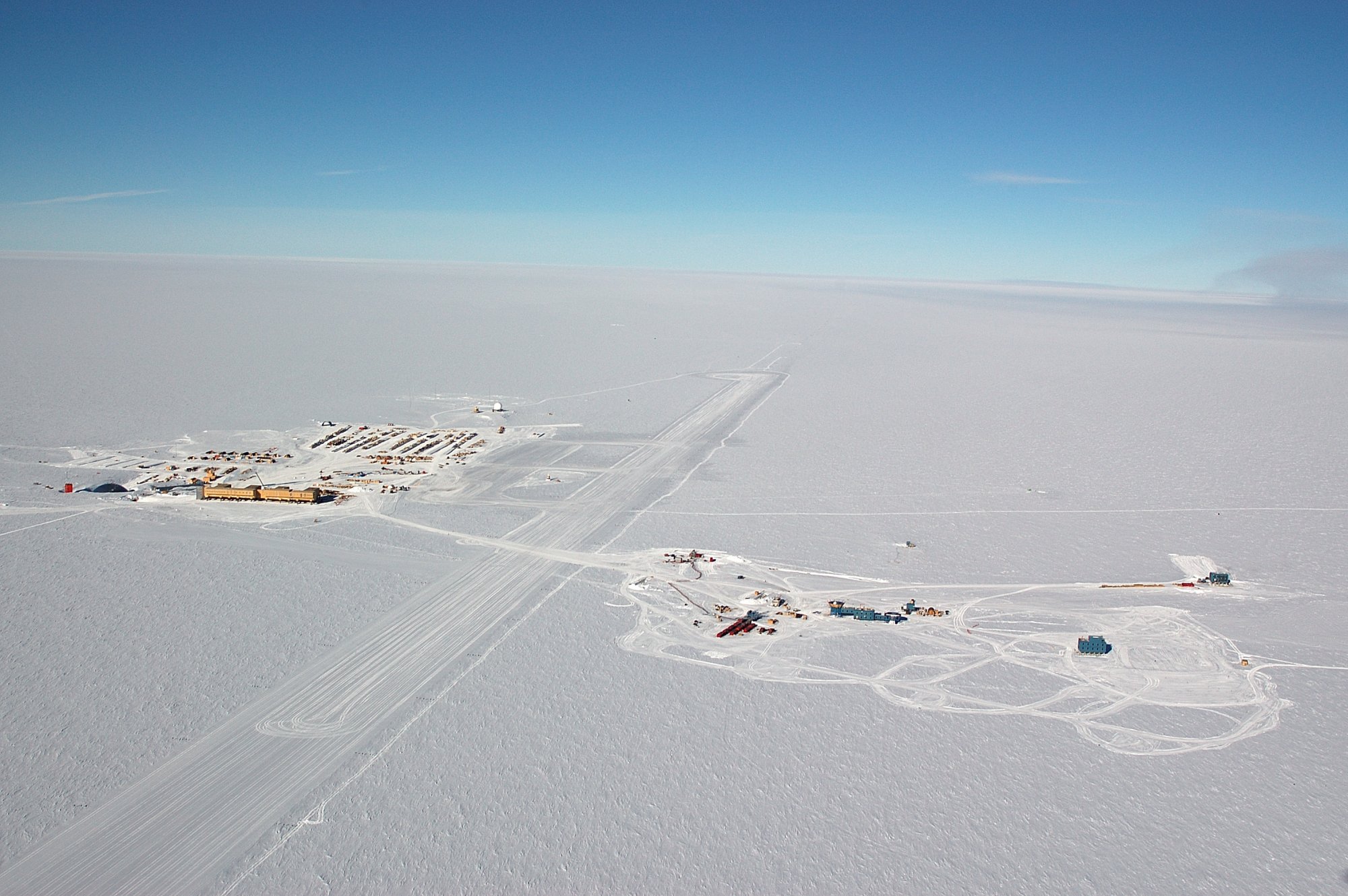 Vue aérienne de la base antarctique Amundsen-Scott, située exactement au pôle Sud géographique. Les lieux de vie et de travail sont à gauche, les grandes expériences dédiées à l'astrophysique et à la cosmologie comme IceCube, Bicep, SPT et Keck Array sont à droite, à environ un kilomètre. On peut se laisser aller à rêver que la base soit la concrétisation de l'Expédition polaire internationale (EPI), la base en Antarctique du roman La nuit des temps de René Barjavel. © Forest Banks, NSF