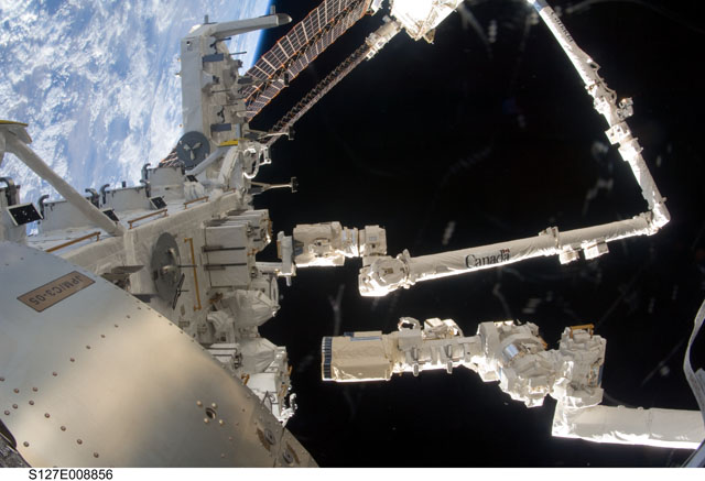 Une partie de la plate-forme des expériences extérieures du module Kibo (Japanese Experiment Module - Exposed Facility). Le Canadarm, bras robotisé de l'ISS, manœuvré ici par la Canadienne Julie Payette, est bien visible. Le plus petit, à droite, est celui de la navette Endeavour. L'image est prise de l'intérieur de Kibo. © Nasa