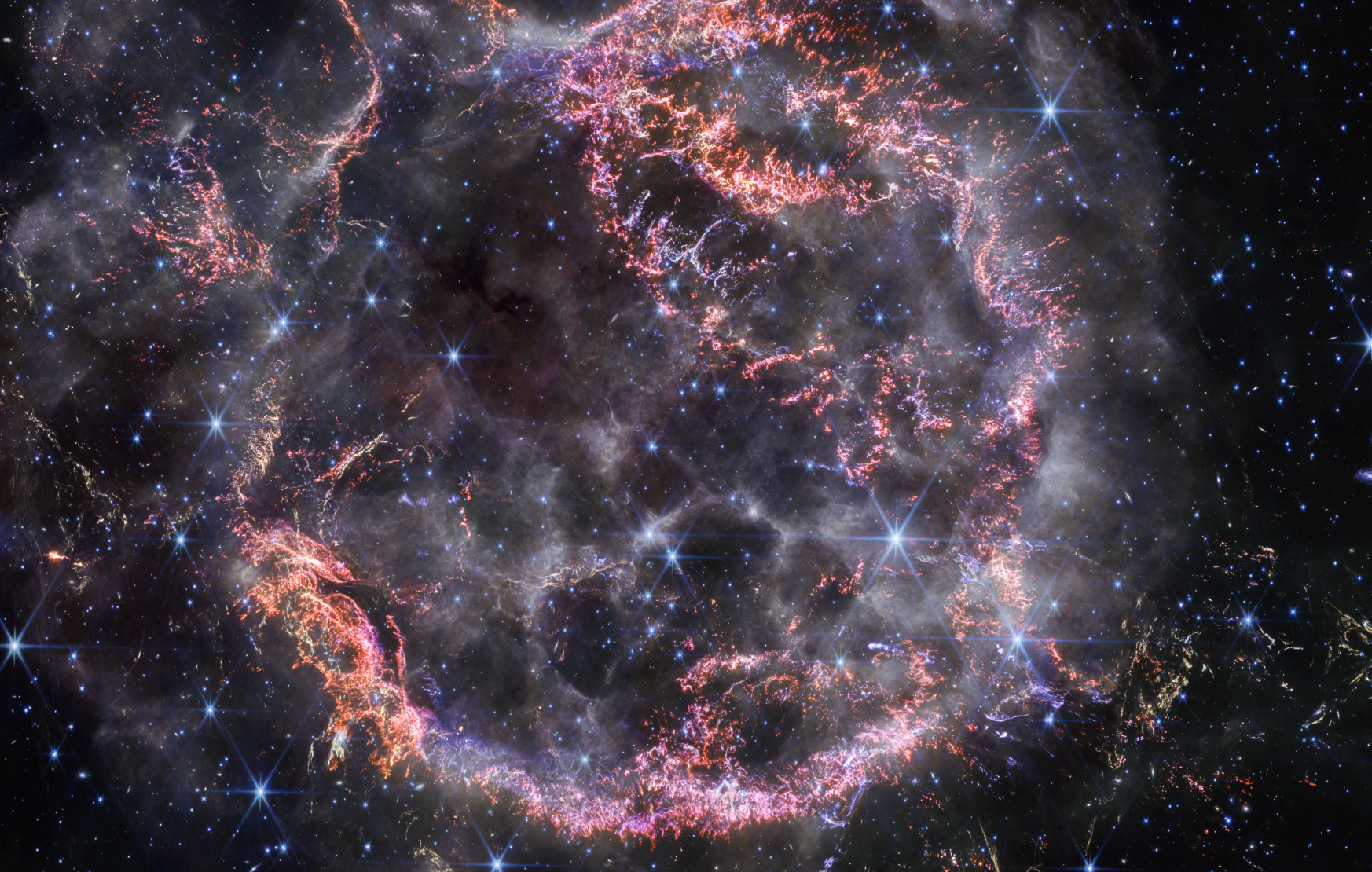 Une nouvelle image haute définition de la NIRCam (Near-Infrared Camera) du télescope spatial James-Webb de la Nasa dévoile des détails complexes du reste de la supernova Cassiopée A (Cas A) et montre la coque de matière en expansion qui heurte le gaz rejeté par l'étoile avant qu'elle n'explose. Les couleurs les plus visibles dans la dernière image de Webb sont des touffes d’orange vif et de rose clair qui constituent la coque interne du reste de la supernova. Ces minuscules nœuds de gaz, composés de soufre, d’oxygène, d’argon et de néon provenant de l’étoile elle-même, ne sont détectables que par la résolution exquise du NIRCam et donnent aux chercheurs une idée de la façon dont l’étoile mourante s’est brisée comme du verre lorsqu’elle a explosé.La périphérie de la coque intérieure principale ressemble à la fumée d’un feu de camp. Cela marque l'endroit où le matériau éjecté de l'étoile explosée s'enfonce dans le matériau circumstellaire environnant. Les chercheurs affirment que cette couleur blanche est la lumière du rayonnement synchrotron, généré par des particules chargées se déplaçant à des vitesses extrêmement élevées en spirale autour des lignes de champ magnétique.Plusieurs échos lumineux sont également visibles sur cette image, notamment dans le coin inférieur droit. C’est l'endroit que la lumière de l’explosion de l’étoile a atteint il y a longtemps, et ce sont des chauffées poussières qui y brillent en se refroidissant. © Nasa, ESA, CSA, STScI, Danny Milisavljevic (Purdue University), Ilse De Looze (UGent), Tea Temim (Princeton University)