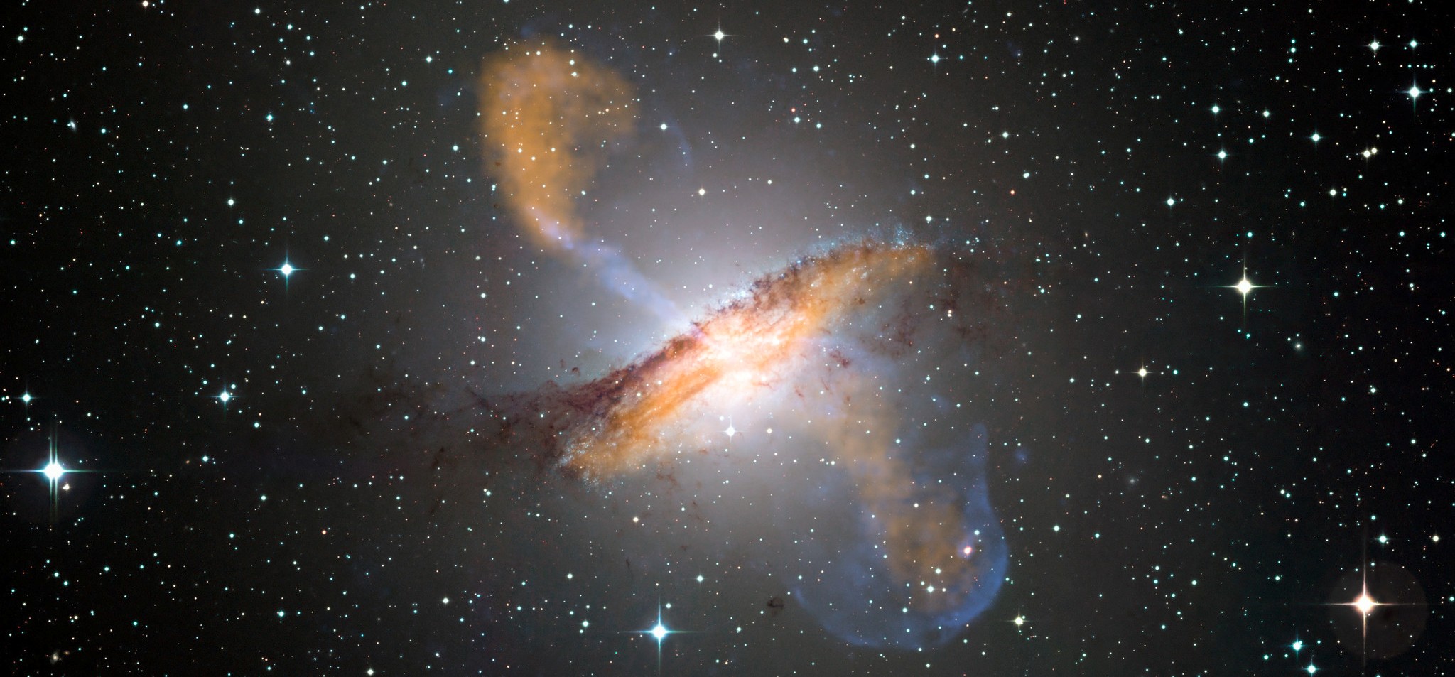 Ces galaxies naines désobéissent-elles à la loi de la gravitation ? Ici, une image en couleurs de Centaurus A, révélant les jets émis par le très actif trou noir central de la galaxie. Ceci est une image composite obtenue avec trois instruments différents fonctionnant à différentes longueurs d'onde. Les données à 870 micromètres, de Laboc, sur Apex, sont montrées en orange. Les données aux rayons X, de l'observatoire&nbsp;de Chandra, sont montrées en bleu. Les données de la lumière visible, du Wide Field Imager (WFI) du télescope MGP/ESO de 2,2 mètres, situé à La Silla (Chili), montre les étoiles. La ligne de poussière caractéristique de la galaxie est proche des « couleurs réelles ». © ESO/WFI (Optical) ; MPIfR/ESO/APEX/A.Weiss et al. (Submillimetre) ; NASA/CXC/CfA/R.Kraft et al. (X-ray)