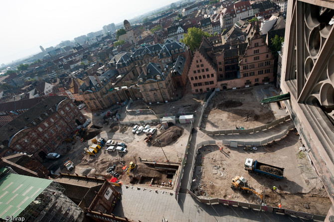 Vue panoramique des bâtiments entourant la place du Château à Strasbourg, en 2012. Le cliché est pris depuis la cathédrale, vers le sud. Au premier plan, l'opération archéologique ; à l'arrière-plan, sur la gauche, l'avancée d'un suivi de réseau. © Inrap