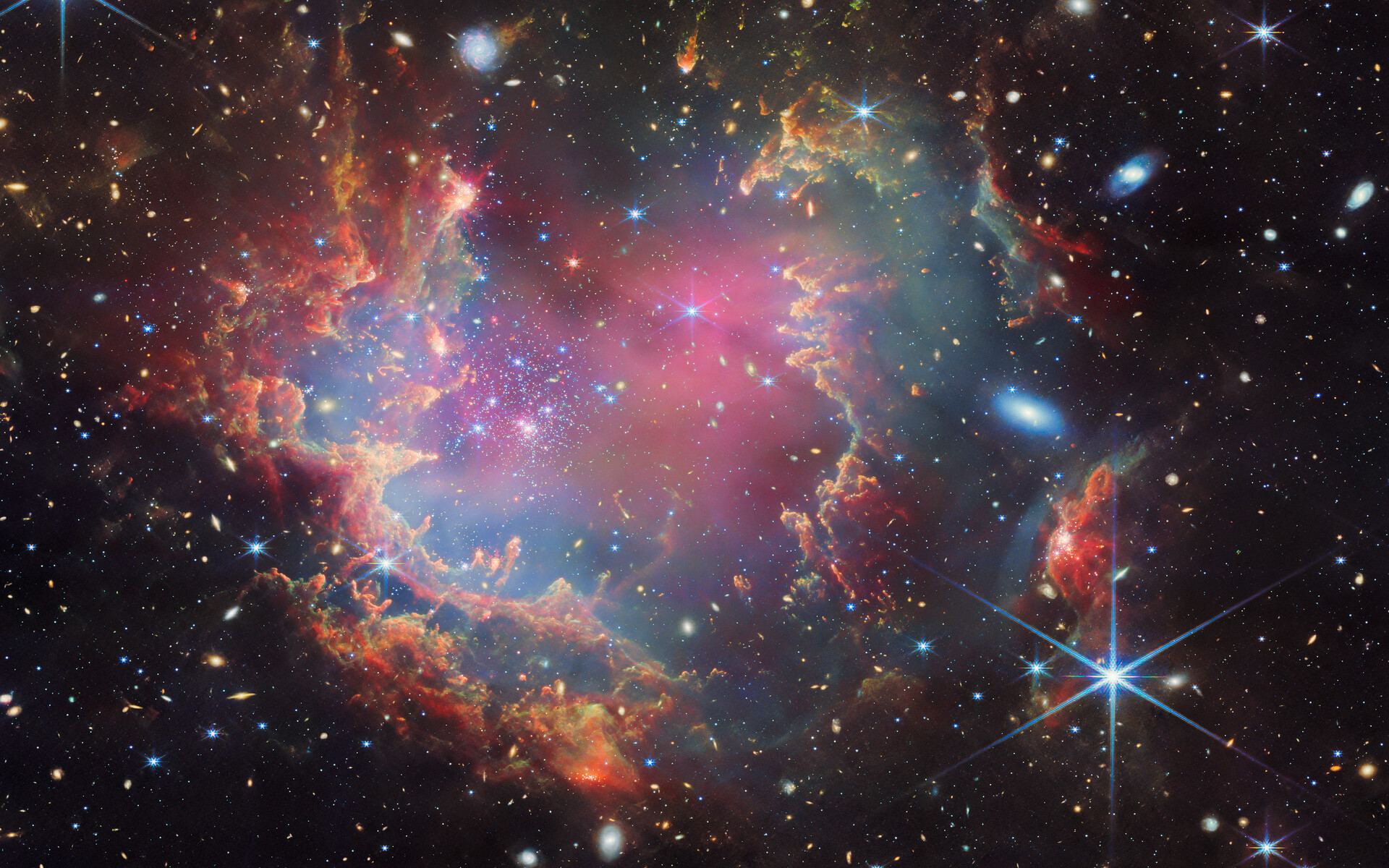 À proximité du Petit Nuage de Magellan, une galaxie située à environ 200 000 années-lumière de la Terre, se trouve le jeune amas d’étoiles NGC 602, présenté sur cette nouvelle image du télescope spatial James-Webb de la Nasa/ESA/CSA. Cette image inclut des données de la NIRCam (Near-InfraRed Camera) et de l’instrument Miri (Mid-InfraRed Instrument) de Webb. Un amas d'étoiles est représenté à l'intérieur d'une grande nébuleuse de gaz et de poussière multicolores. La matière forme des crêtes et des pics sombres de gaz et de poussière entourant l'amas, éclairés sur le côté intérieur, tandis que des couches de nuages ​​diffus et translucides les recouvrent. Autour et à l'intérieur du gaz, on peut voir un grand nombre de galaxies lointaines, certaines assez grandes, ainsi que quelques étoiles plus proches de nous qui sont très grandes et brillantes. © ESA/Webb, Nasa &amp; CSA, P. Zeidler, E. Sabbi, A. Nota, M. Zamani (ESA/Webb)